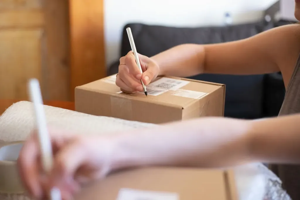 Hands writing an address label on a cardboard parcel, the kind of order an AI agent for eBay sellers must leave a human to check