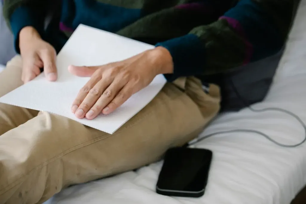 Person reading a braille page with a smartphone resting on the bed beside them, showing AI agents for accessibility alongside traditional tools
