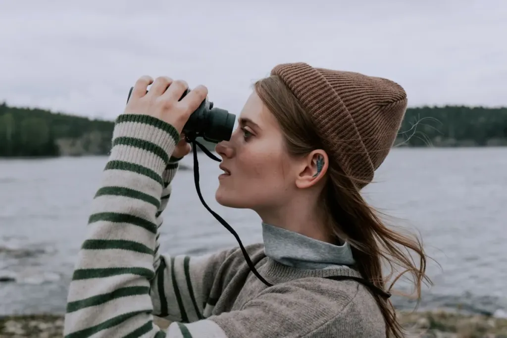 Woman in a knitted hat wearing a hearing aid while looking through binoculars by a lake